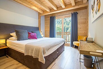 Cozy wooden bedroom with a large bed, colorful pillows, balcony with forest view, and a small desk/chair setup.