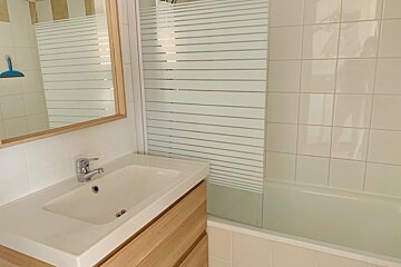 Modern bathroom with a wooden vanity, white sink, mirror, and a tiled shower/tub combo with a frosted glass screen. Neutral colors.