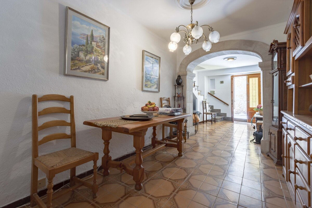 A traditional hallway features intricate tiled floors, a rustic wooden table with chairs, and an archway leading to a bright entrance. Paintings adorn white walls.
