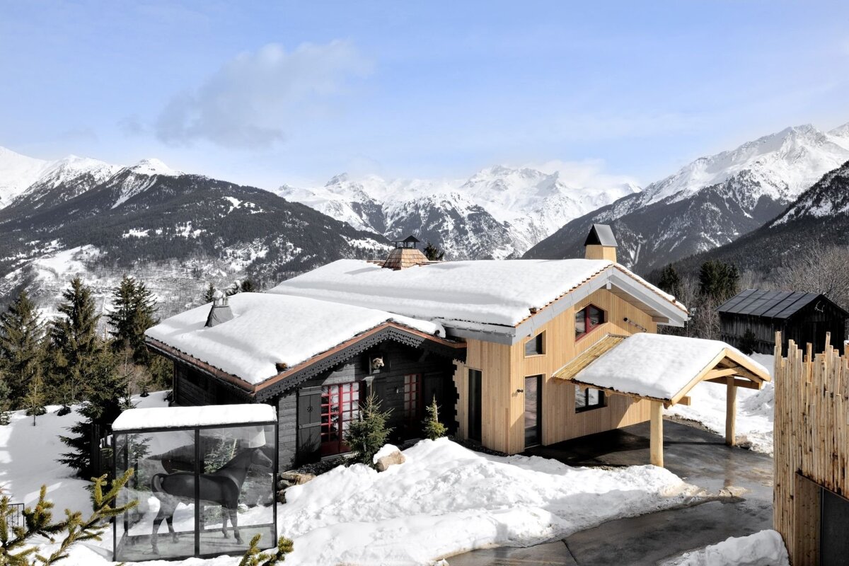 Snow-covered alpine chalet with a horse sculpture in a glass case, set against majestic snowy mountains and pine forests under a blue sky.