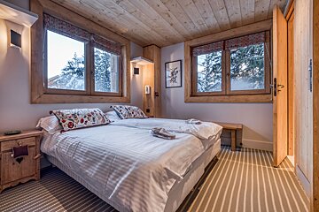 A cozy chalet-style bedroom with twin beds, a wooden ceiling, striped carpet, and two windows overlooking a snowy winter forest.