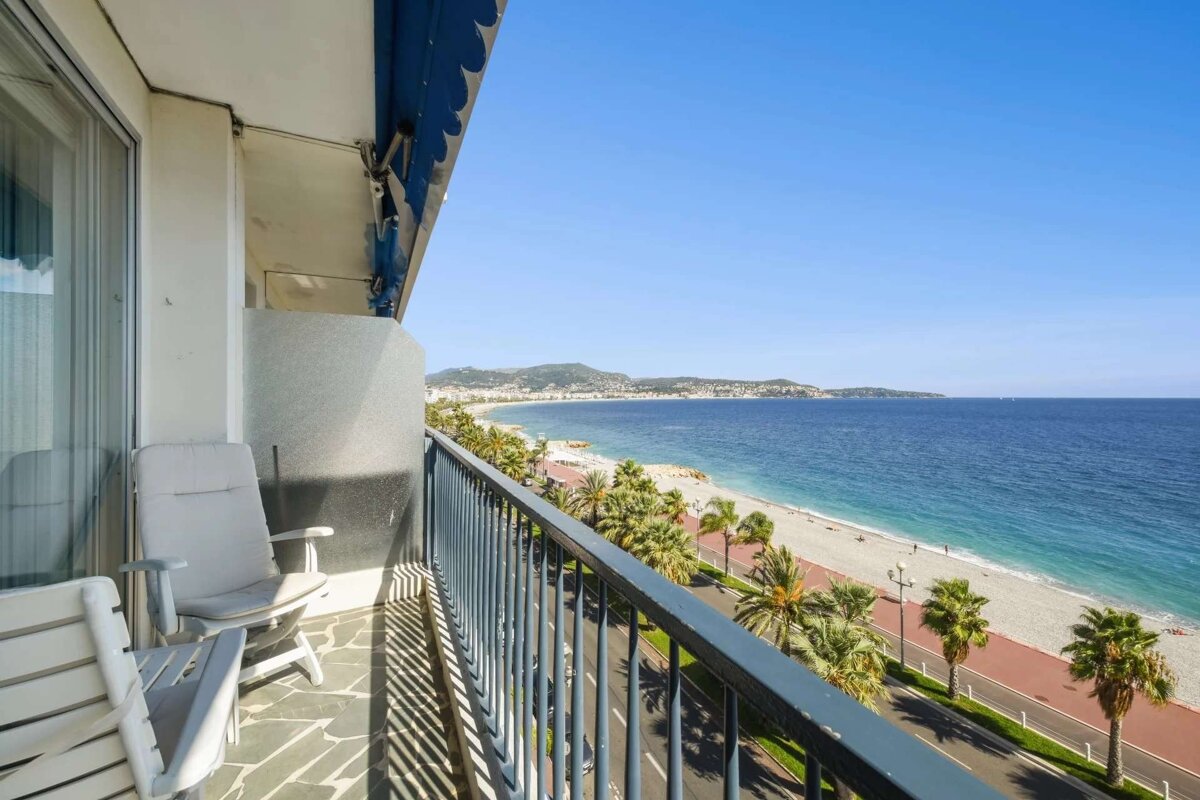 A sunny balcony with chairs overlooks a vibrant beach, palm-lined promenade, and the sparkling blue sea extending to a coastal city under a clear sky.