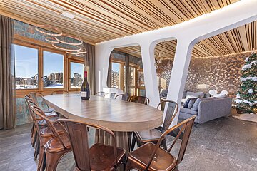 A modern rustic dining room with a long table, metal chairs, and a wine bottle, offering snowy mountain views. Adjacent is a living area with a Christmas tree.