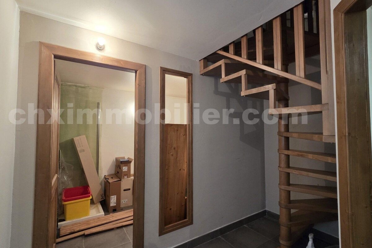 A hallway with a wooden spiral staircase, an open doorway revealing moving boxes, and a narrow wooden panel on neutral walls.