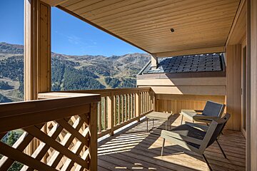 Sunny wooden balcony with outdoor seating overlooks a vast mountain range and forest under a clear blue sky.