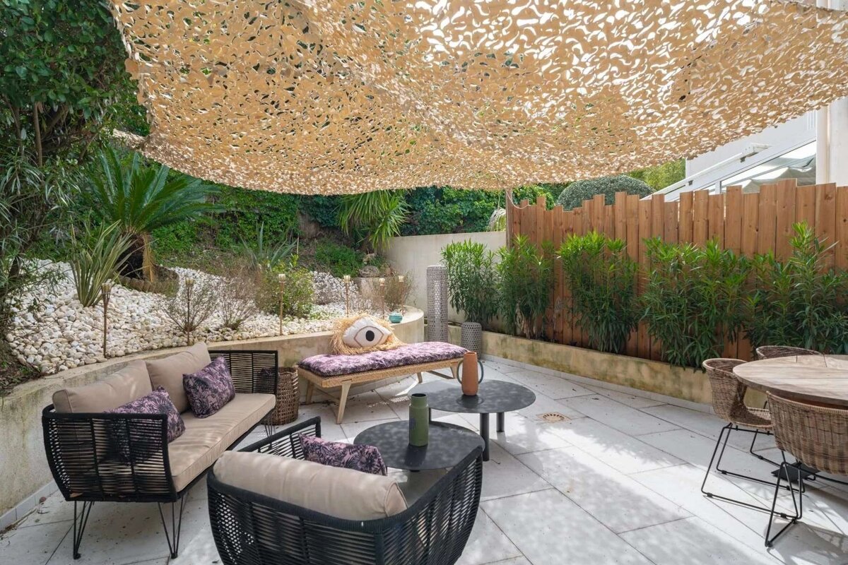 A cozy outdoor patio with modern lounge furniture, shaded by a camouflage net. Lush greenery, white pebbles, and a wooden fence line the space.