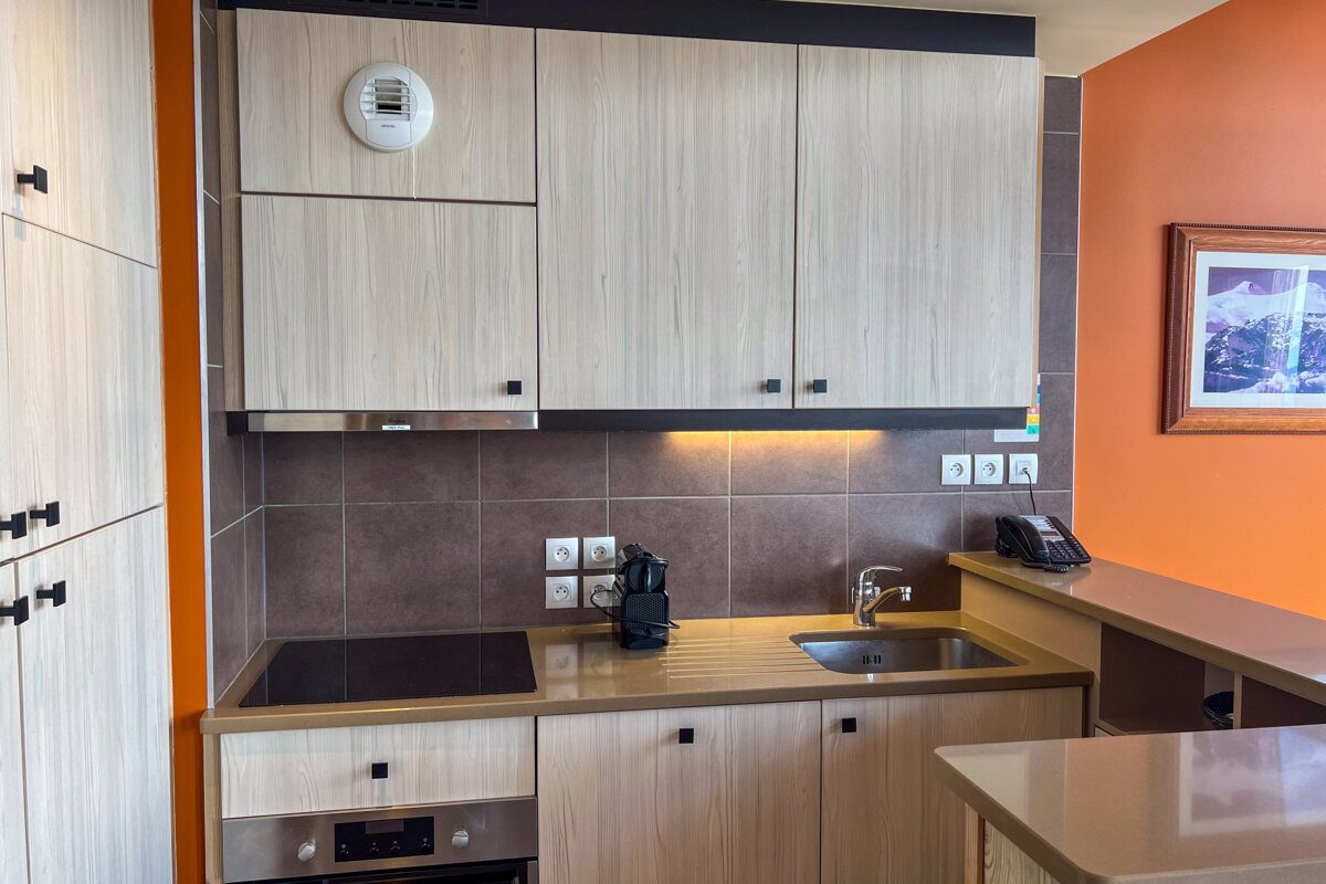 A modern kitchen with light wood cabinets, brown tiled backsplash, and a striking orange accent wall. Includes a hob, sink, oven, and mountain art.