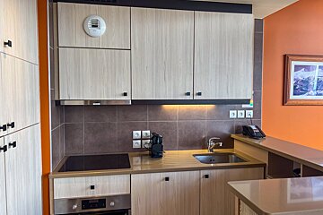 A modern kitchen with light wood cabinets, brown tile backsplash, and an orange wall. It features an induction hob, oven, sink, and telephone.