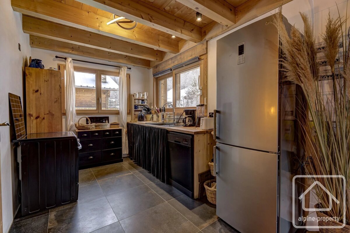Cozy rustic kitchen with exposed wooden ceiling beams, dark tiled floor, stainless steel refrigerator, and wooden cabinetry.