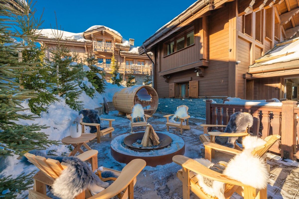 Snowy chalet patio with fire pit, wooden chairs with fur throws, and a barrel sauna, surrounded by pine trees under a blue sky.