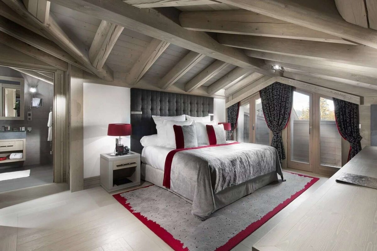 A luxurious chalet-style bedroom with exposed wooden beams, a gray bed with red accents, French doors, and a modern attached bathroom.