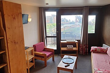 A snug studio room featuring wooden furniture like a red armchair, daybed, and shelves. Big windows overlook a village nestled in green hills.