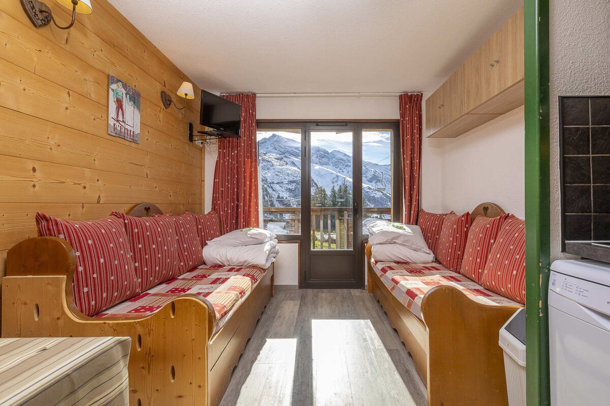 Cozy ski chalet room with two daybeds, red cushions, wood-paneled walls, and a large window framing a beautiful snowy mountain landscape.