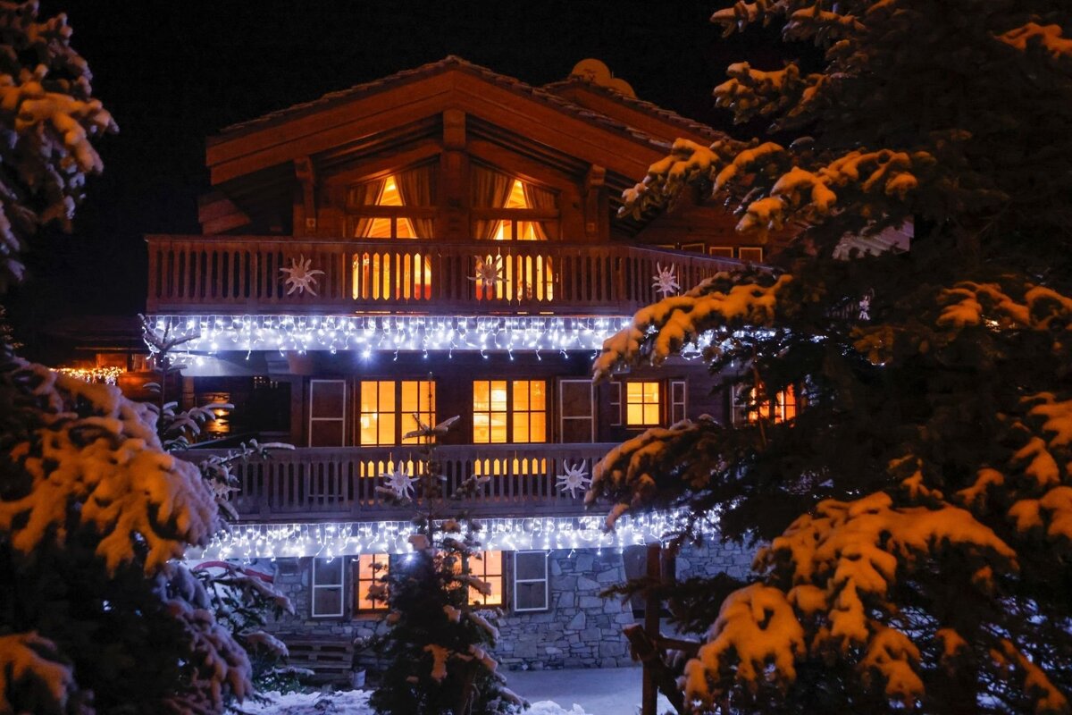 A cozy wooden chalet at night, adorned with icicle lights. Snow covers pine trees and the ground, and warm light glows from the windows.