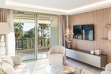 Bright, modern living room with neutral decor, featuring a large balcony overlooking palm trees and the ocean. Includes a wall-mounted TV and elegant seating.