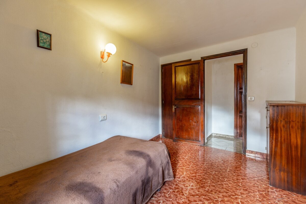 A simple bedroom with a single bed and brown blanket, white walls, patterned red-brown tiled floor, and dark wooden doors, with a wall light.