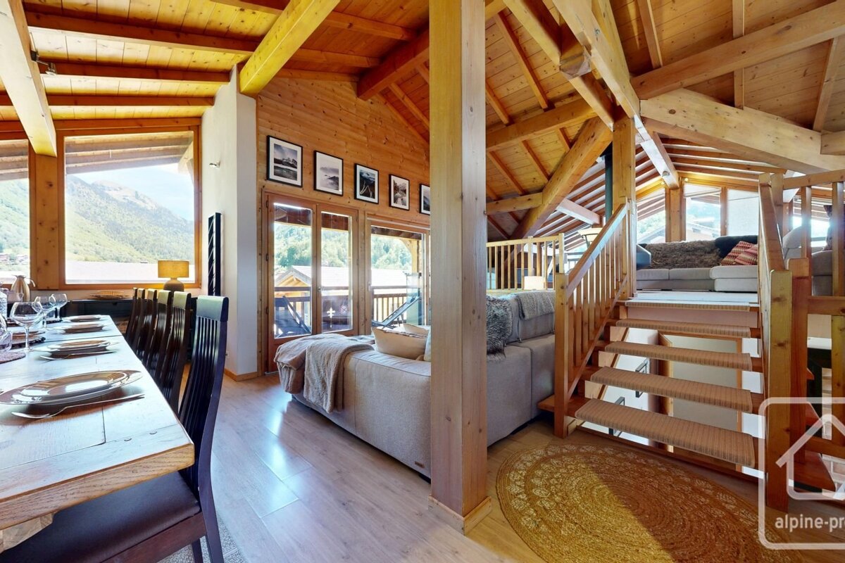 An interior of a wooden cabin with a living room, dining room, and staircase, with a scenic view of mountains outside.