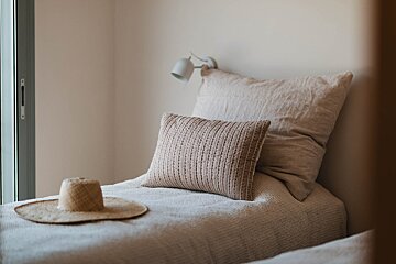 A neutral-toned bedroom featuring a bed with linen pillows, a quilted pillow, and a straw hat, creating a serene, minimalist ambiance.