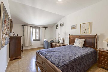 Cozy bedroom with a traditional wooden bed, blue patterned duvet, tiled floor, white walls, and a window with white curtains. Wooden furniture and wall art complete the space.