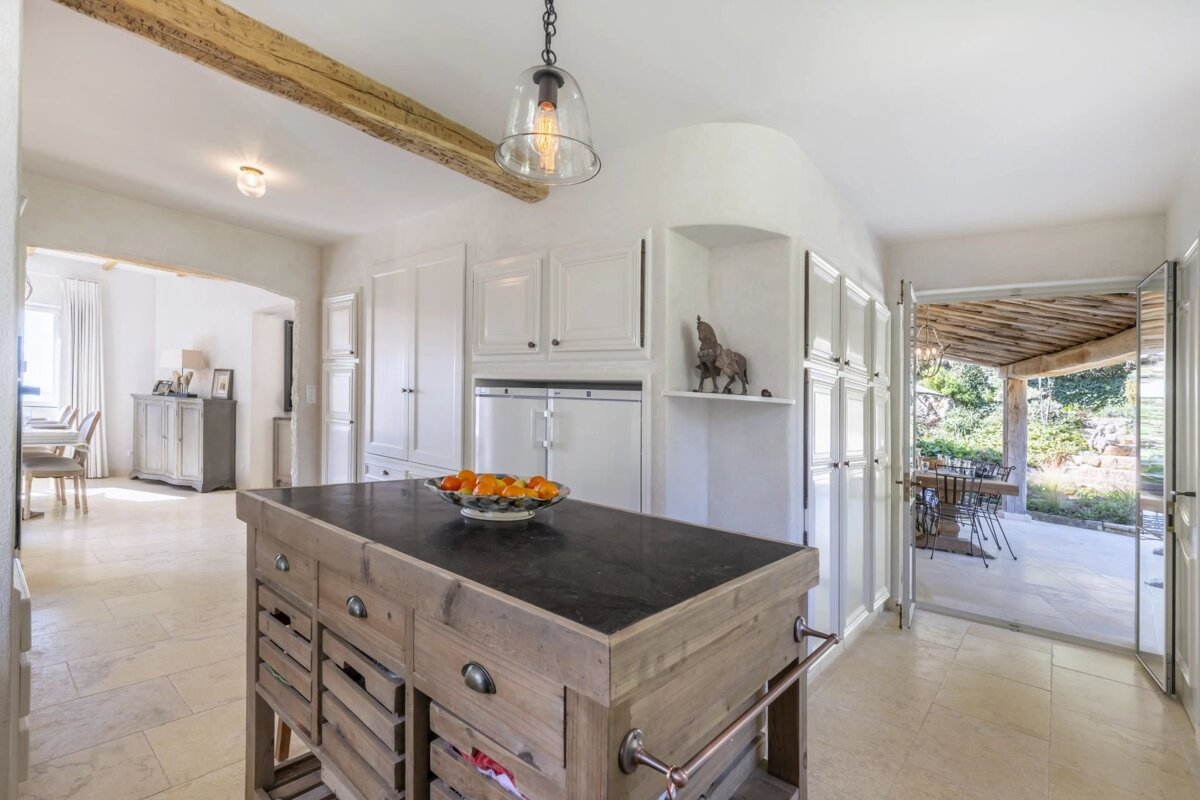 A rustic kitchen featuring a wooden island with a bowl of oranges, white cabinetry, an exposed beam, and an open view to an outdoor patio.