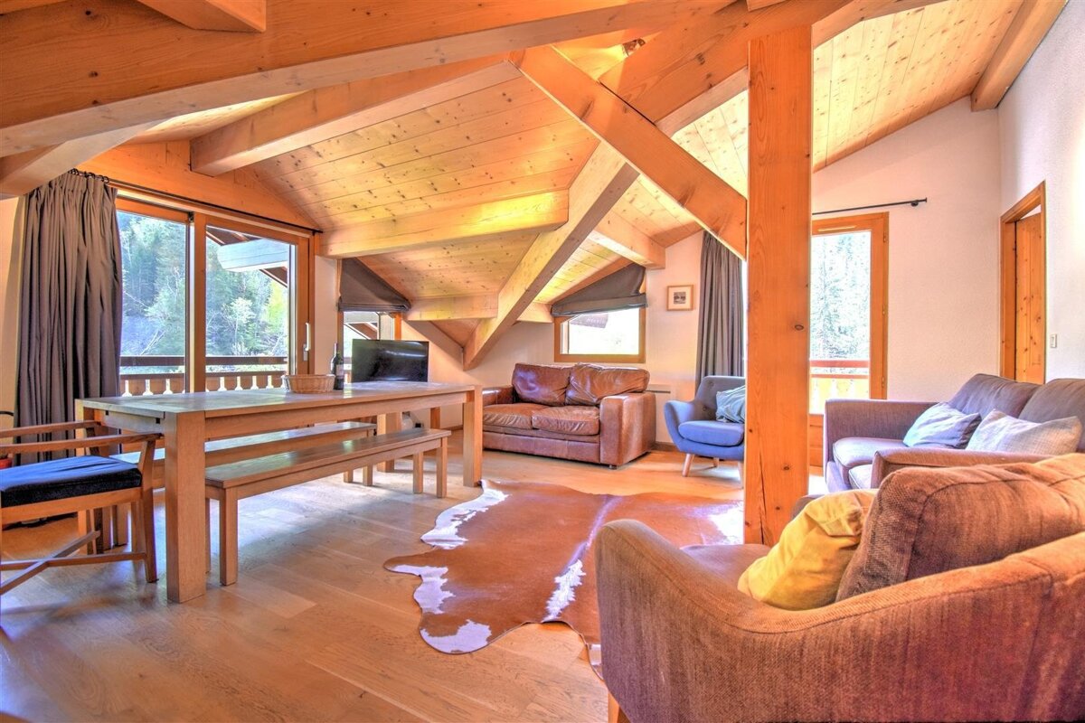 Cozy chalet living area with rustic wood beams, a dining table, comfy sofas, a cowhide rug, and large windows overlooking a forest.