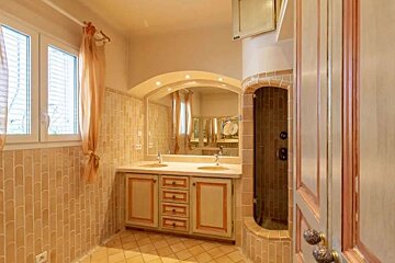 Warm-toned bathroom with double vanity, large mirror, and tiled shower alcove. A window with peach curtains adds natural light.