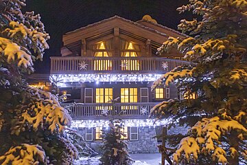 A snow-covered wooden chalet illuminated with warm window lights and festive icicle string lights at night, framed by snowy pine trees.