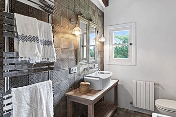 A well-lit bathroom featuring patterned tiles, a wooden vanity with a sink, a towel warmer, and a window with a green outdoor view.
