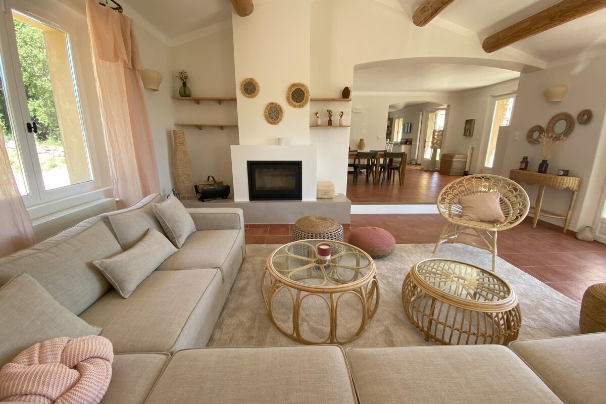 A bright, airy living room with a neutral sectional, rattan furniture, fireplace, and exposed beams. Terracotta floors extend to a dining area beyond.