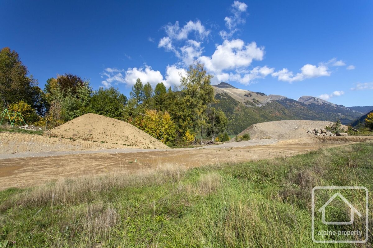 A construction site in a mountain valley, with large dirt piles, green and autumn-colored trees, under a bright blue sky.