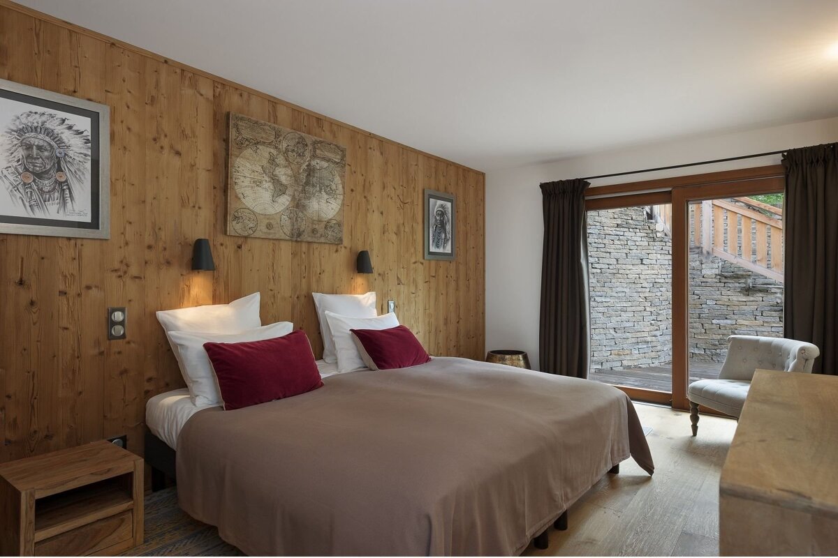 Cozy bedroom with wooden paneled walls, a large bed with red pillows, and art. A sliding glass door reveals an outdoor stone wall and steps.