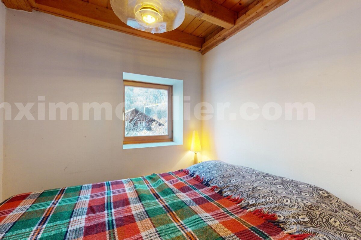 A cozy bedroom with a plaid bed, wooden ceiling, and a window framing a snowy mountain scene with a rustic house.
