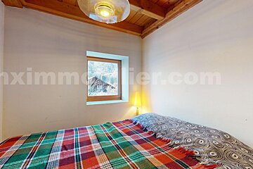 A cozy bedroom with a plaid bed, wooden ceiling, and a window framing a snowy mountain scene with a rustic house.
