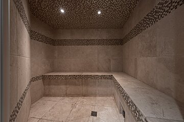 A neutral-toned, tiled steam room with a built-in bench. Mosaic bands accent the walls and ceiling, which also features two recessed lights.