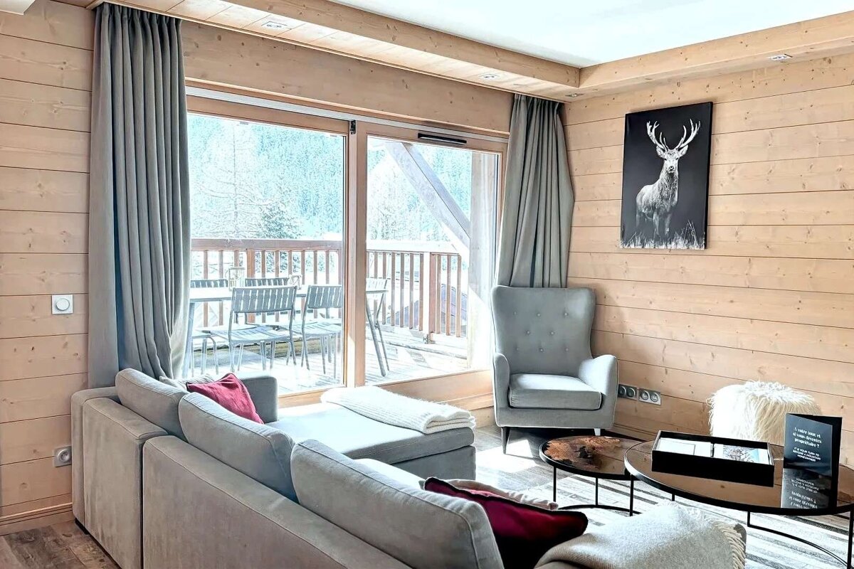 A warm, rustic living room featuring wood-paneled walls, a gray sofa and armchair, deer artwork, and a balcony overlooking a snowy forest.