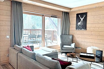 A warm, rustic living room featuring wood-paneled walls, a gray sofa and armchair, deer artwork, and a balcony overlooking a snowy forest.