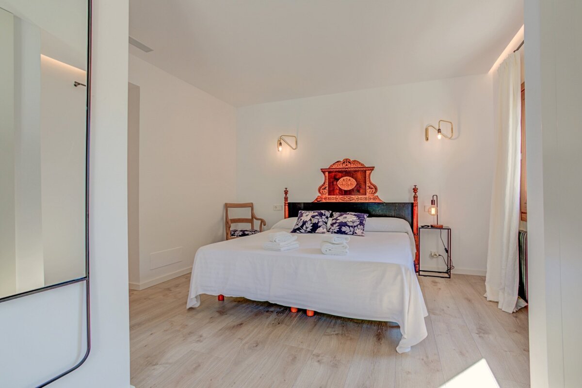 A bright, white bedroom featuring a striking antique wooden headboard, white bed linen, and light wooden floors. Modern minimalist with charm.