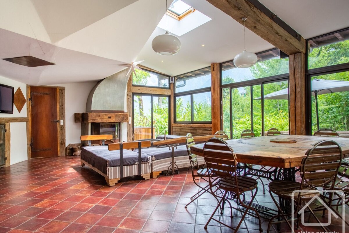 A rustic, airy living room with large windows framing lush green views, exposed beams, a cozy fireplace, and a communal dining area.