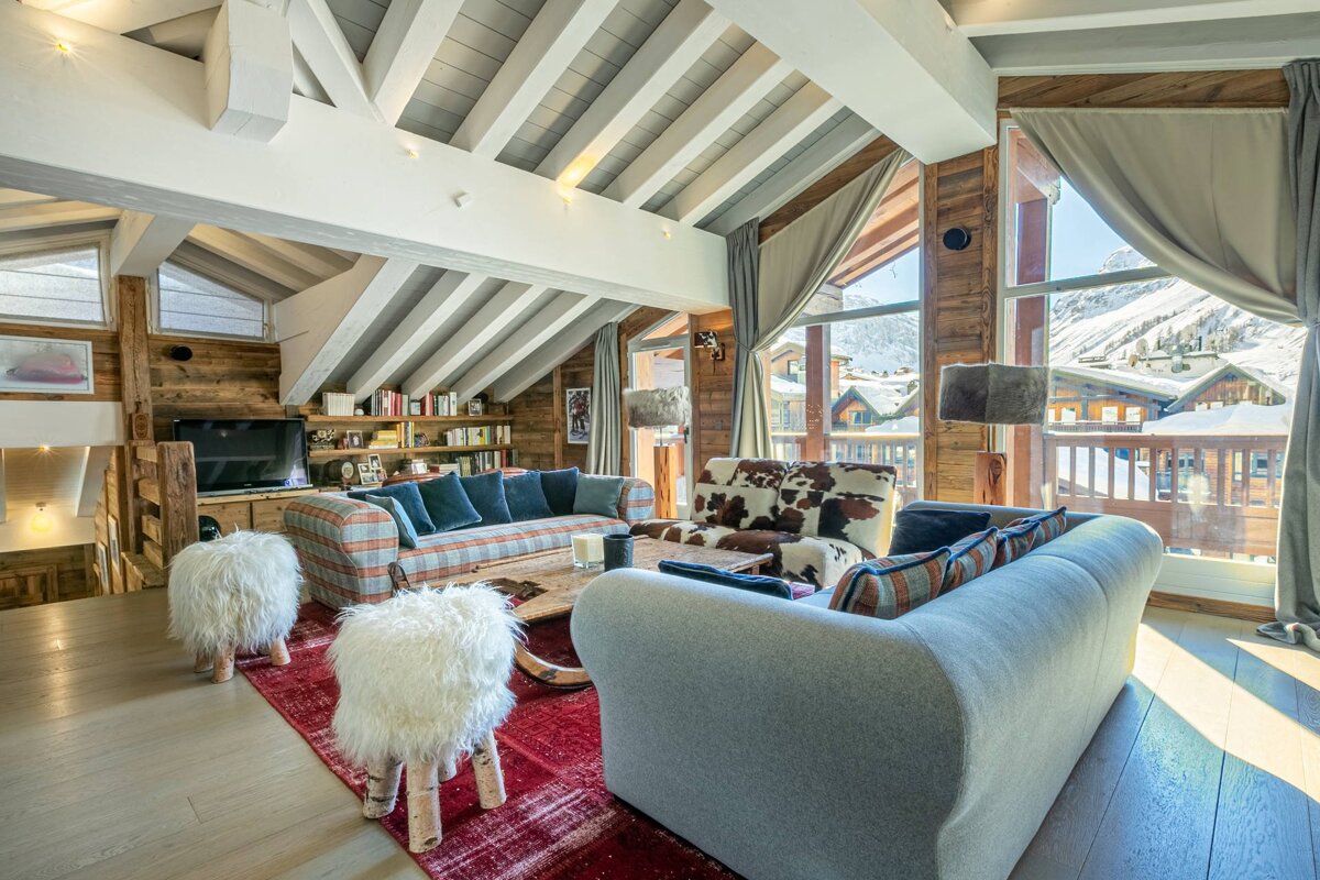A cozy chalet living room with vaulted wood-beamed ceilings, plush seating, and large windows overlooking a snowy mountain resort.