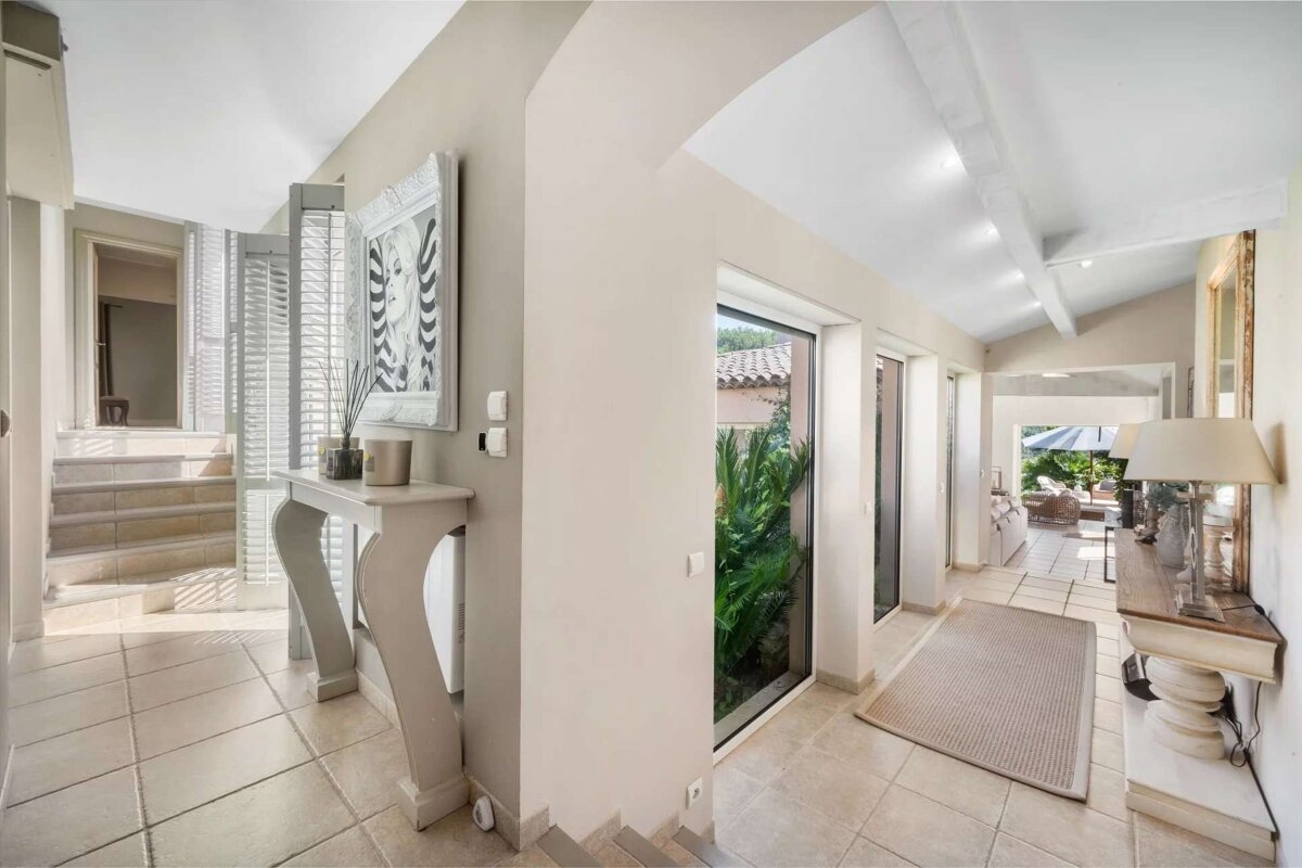 A bright, neutral-toned interior hallway features tiled floors, a console table with decor, and large windows looking onto lush greenery. A staircase ascends on the left, while the hall opens to anoth