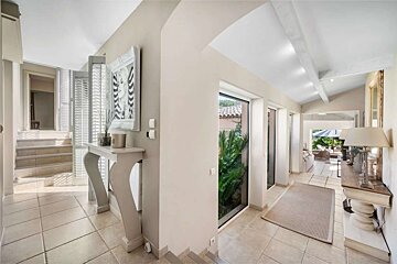 A bright, neutral-toned interior hallway features tiled floors, a console table with decor, and large windows looking onto lush greenery. A staircase ascends on the left, while the hall opens to anoth