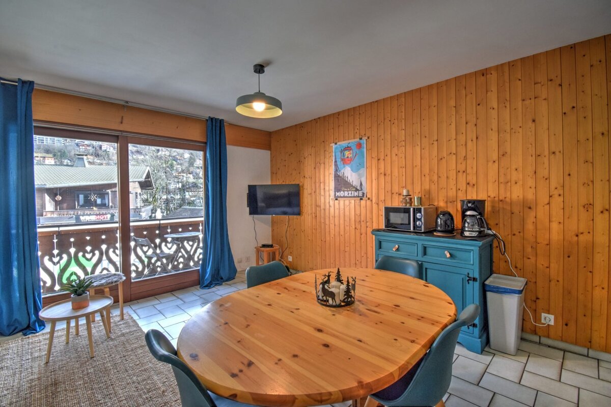 A warm, wood-paneled dining area with an oval table, blue chairs, and a balcony offering mountain village views. Features a small kitchen setup.