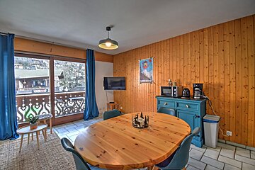 A warm, wood-paneled dining area with an oval table, blue chairs, and a balcony offering mountain village views. Features a small kitchen setup.