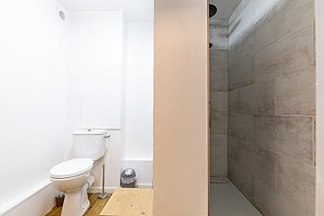 A bright bathroom features a white toilet on the left and a tiled walk-in shower on the right, separated by a light-colored divider wall.