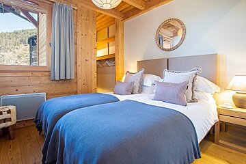 Cozy chalet bedroom featuring two beds with blue blankets, wooden paneling, a round mirror, and a window looking out at mountains.