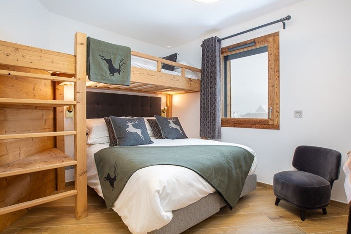 Cozy bedroom featuring a wooden bunk bed over a double bed with deer-themed decor, a grey armchair, and a window.