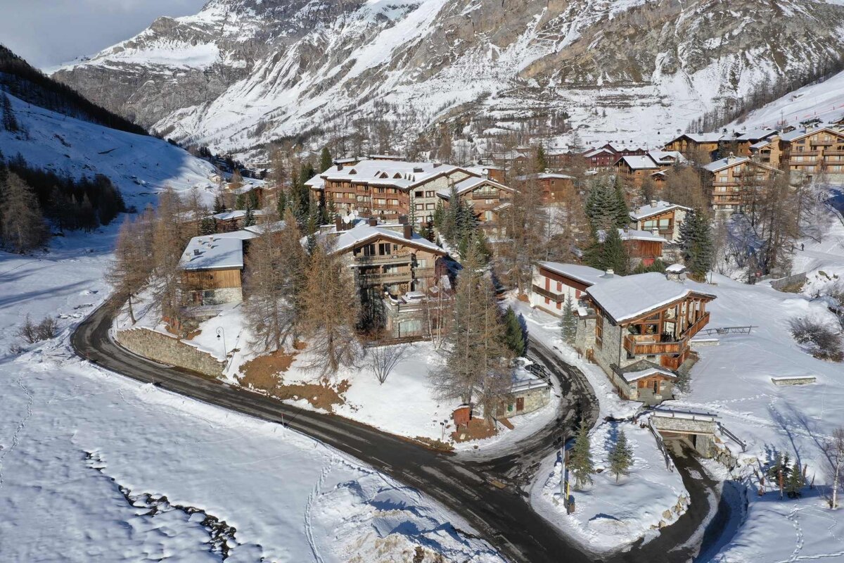 Aerial view of a snowy alpine village with traditional chalets, winding roads, and majestic snow-capped mountains in a winter valley.