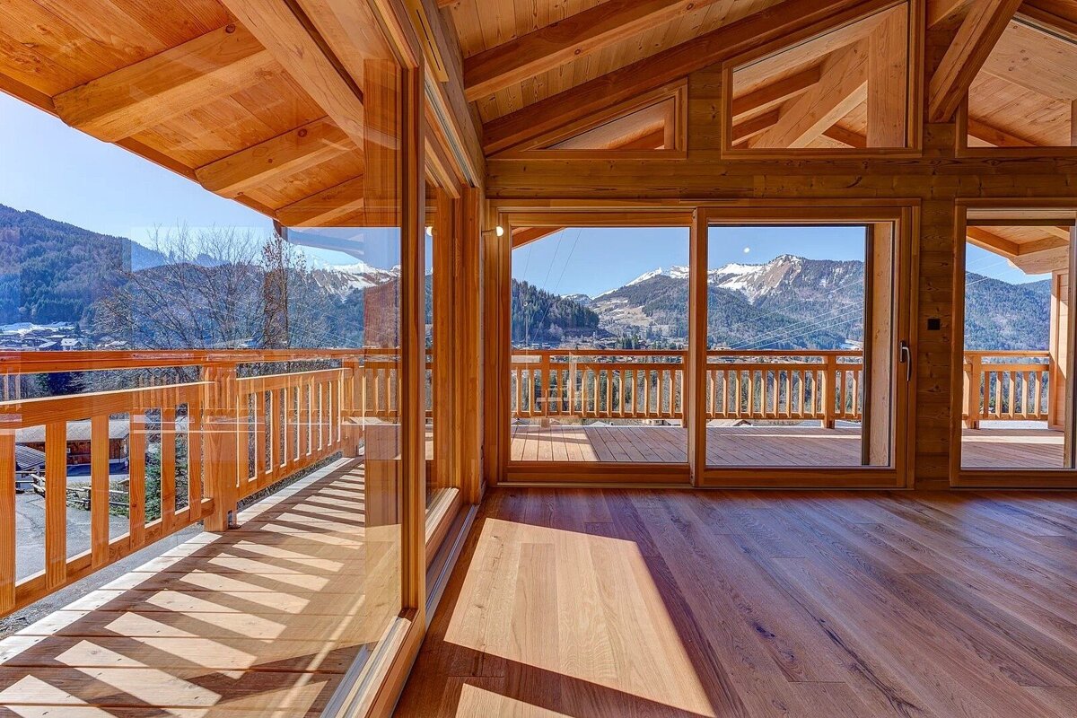Bright wooden cabin interior with large glass doors leading to a balcony with spectacular views of snow-capped mountains.
