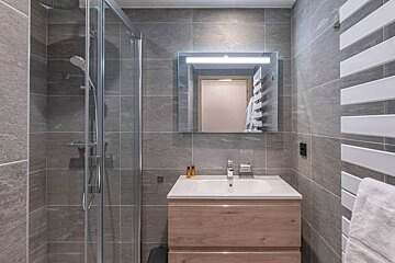 Modern bathroom with grey tiled walls, a glass shower cabin, a wooden vanity, and an illuminated mirror.