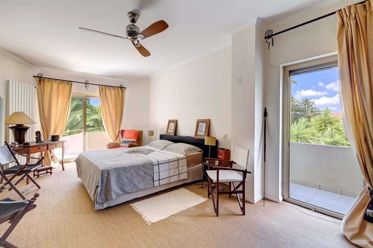 A bright bedroom with a bed, ceiling fan, desk, and balcony offering a lush green view. Beige carpet and warm-toned curtains create a cozy feel.